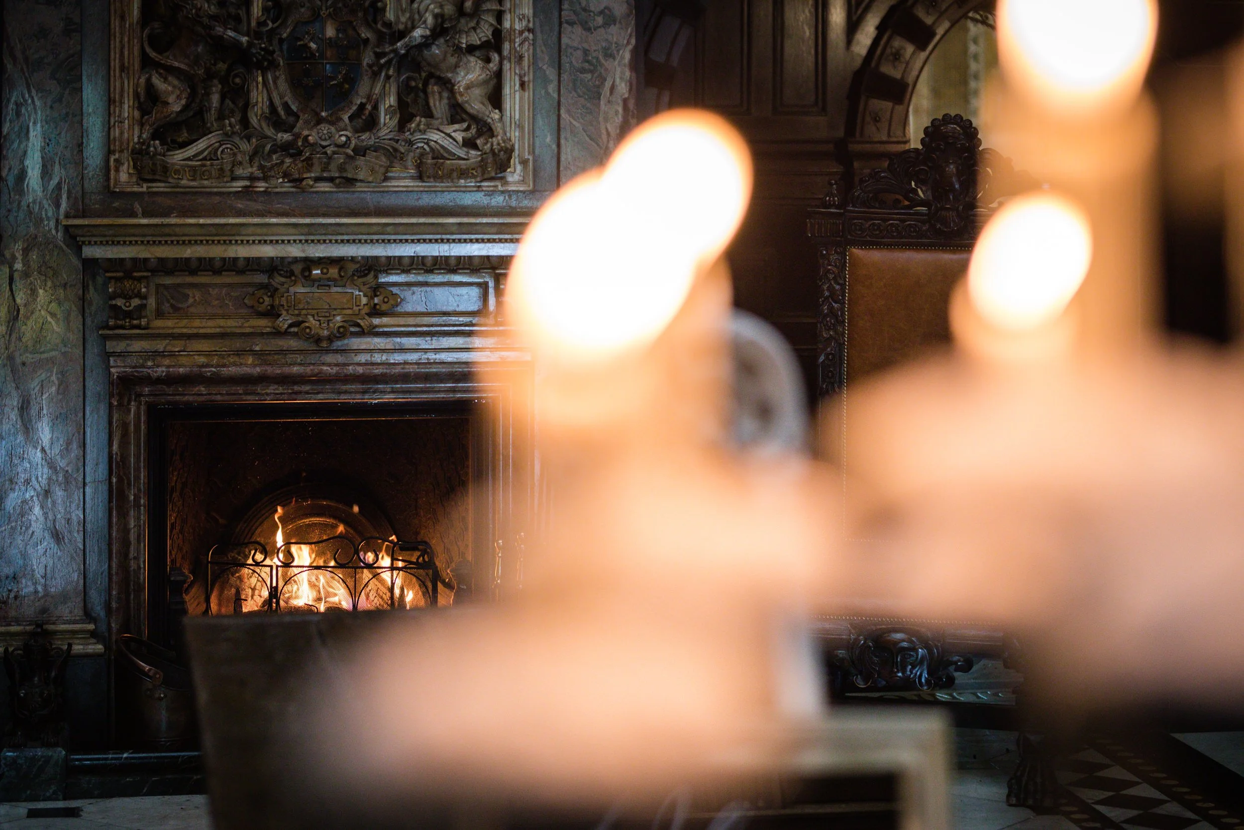 A fireplace with a roaring fire in an ornately decorated room, framed by blurred foreground objects.