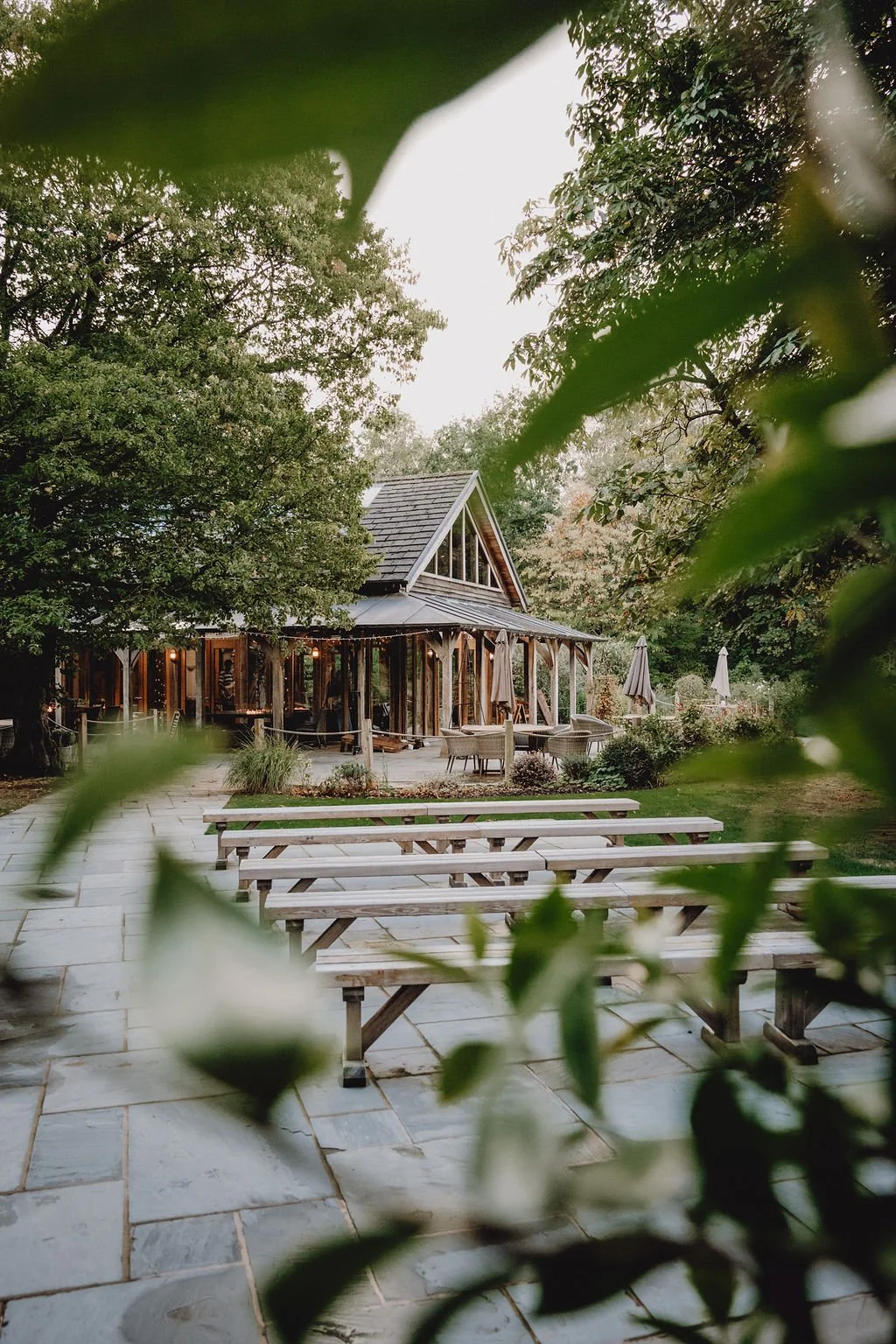 A rustic outdoor gathering space with picnic tables, surrounded by lush green trees, and a wooden building with a gabled roof, large windows, and string lights.