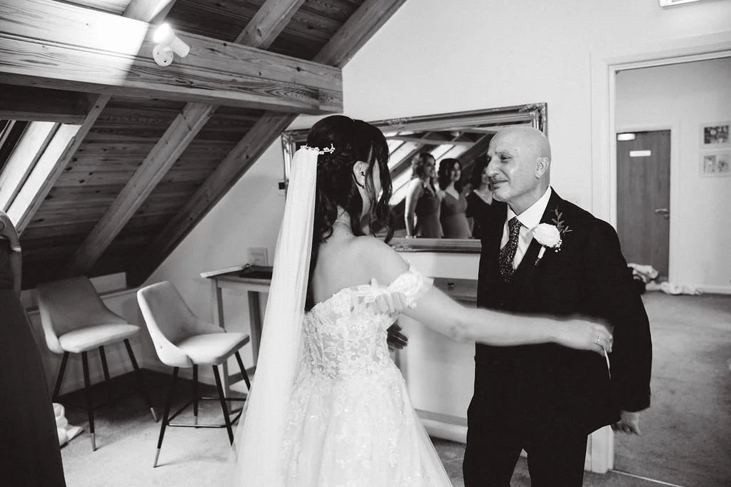 Bride in a wedding dress and veil dancing with an older man in a suit in a room with chairs and a sloped wooden ceiling.