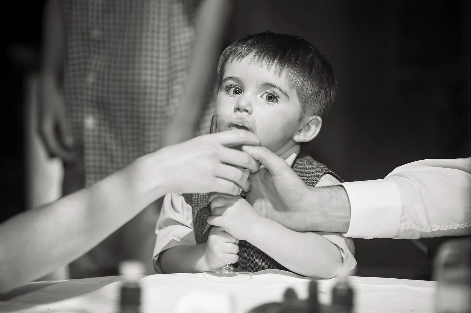Infant boy clings to champagne glass and both parents try to take it away from him