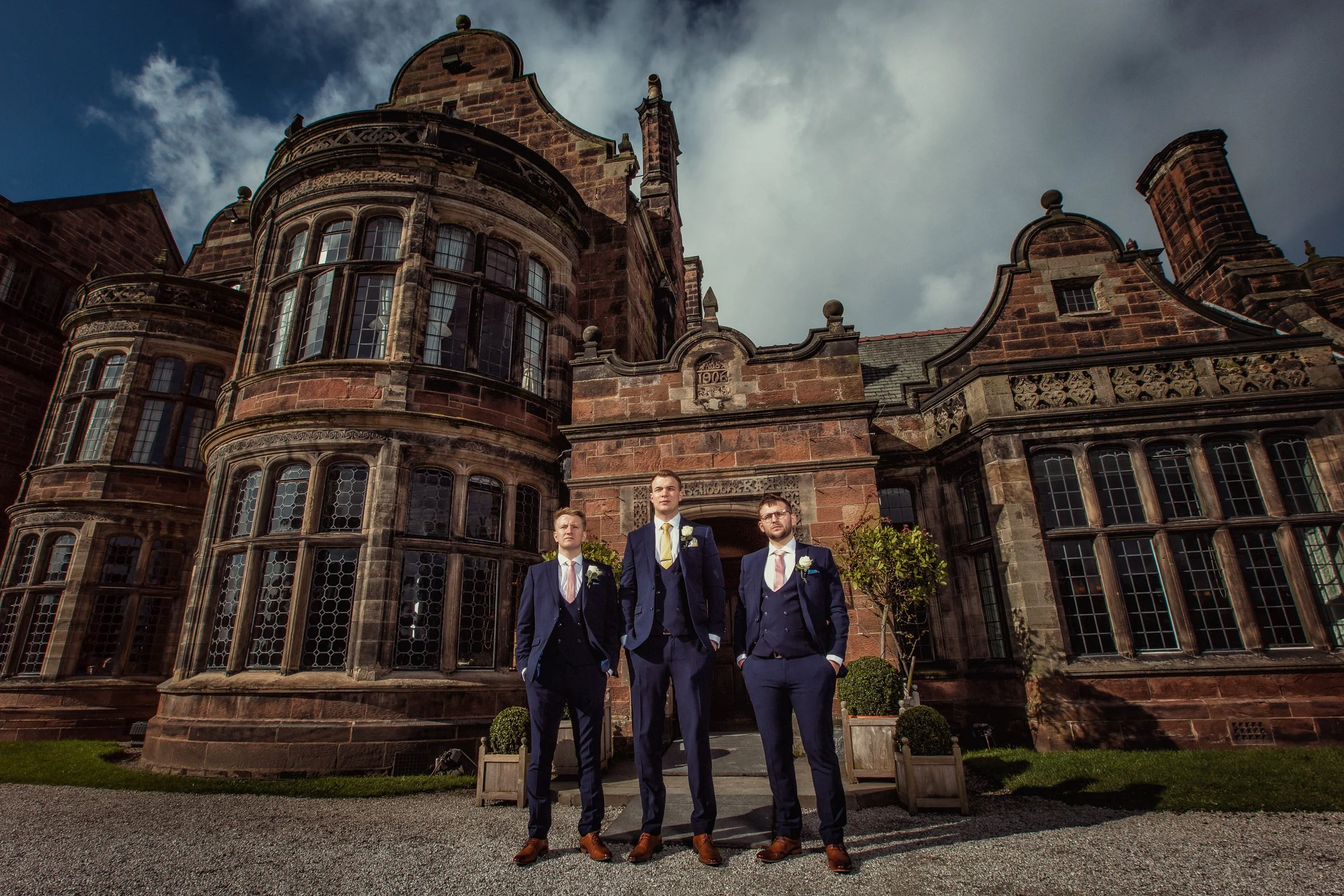 Three men in suits standing in front of a historic brick building, likely a castle or mansion, on a partly cloudy day.