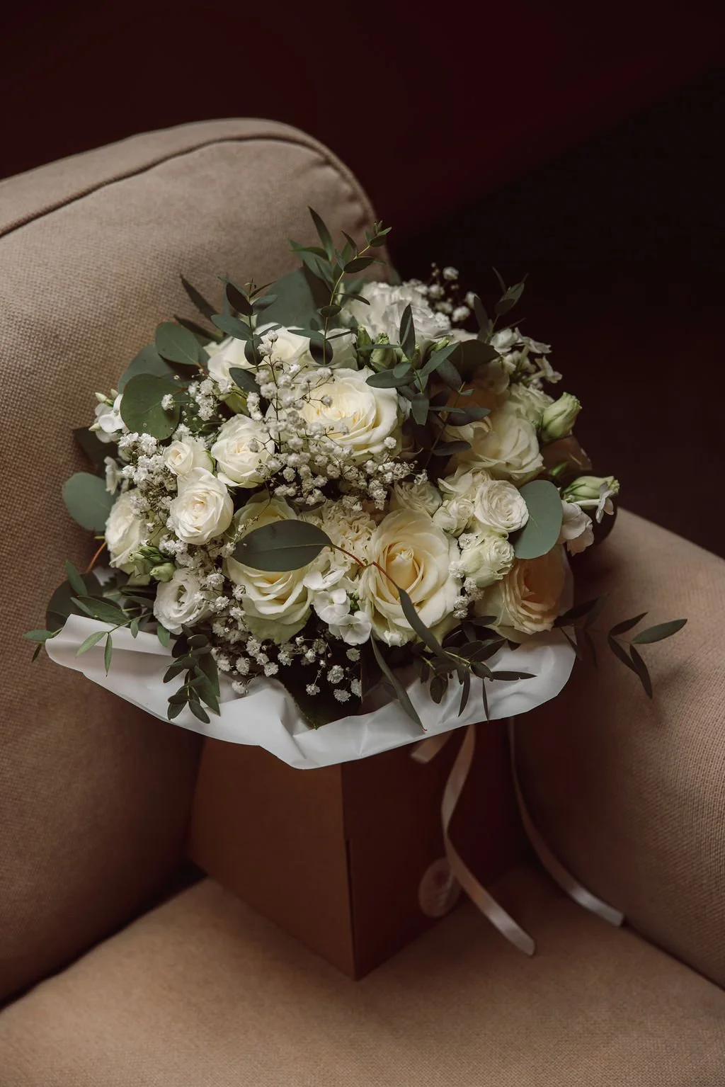 A bouquet of white roses, baby's breath, and greenery resting on a beige upholstered chair with a brown box underneath.