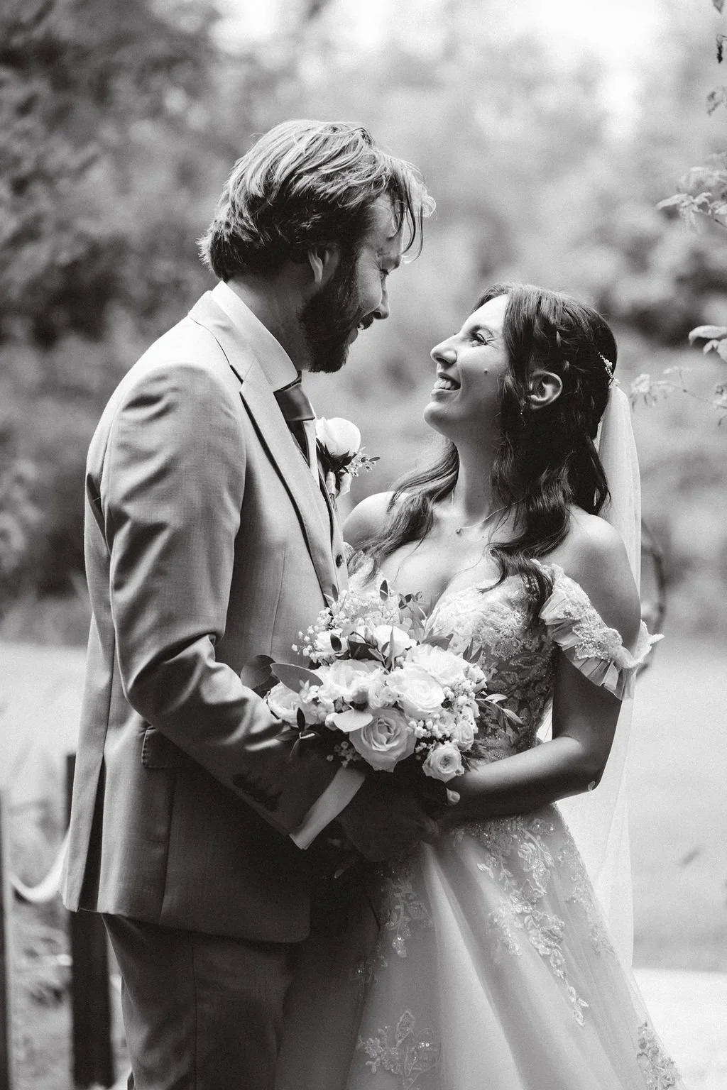 A black and white photo of a couple on their wedding day smiling at each other, with the groom holding a bouquet of flowers.