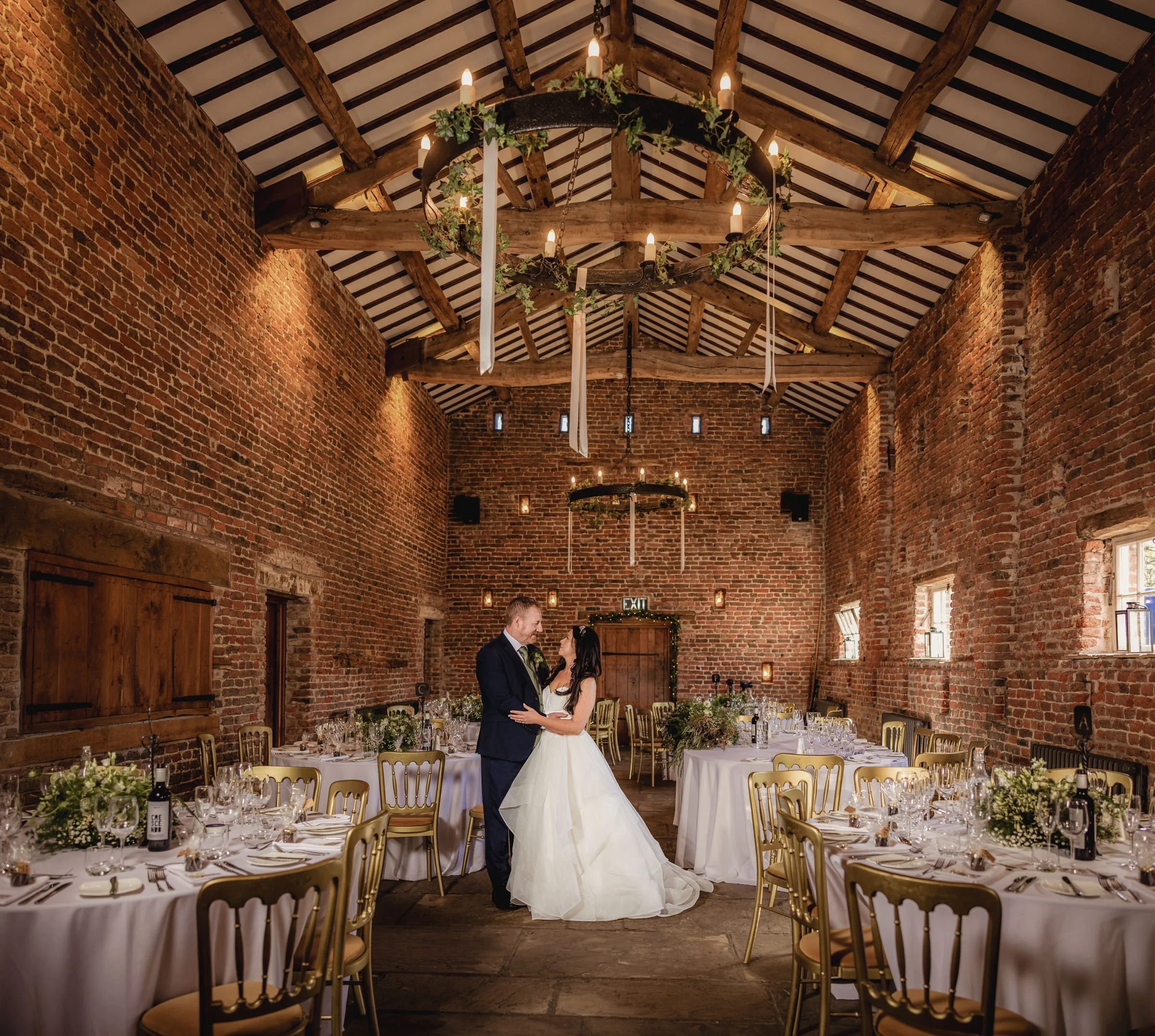 A bride and groom dancing in a rustic wedding reception hall with exposed brick walls and a vaulted ceiling with wooden beams and chandeliers.