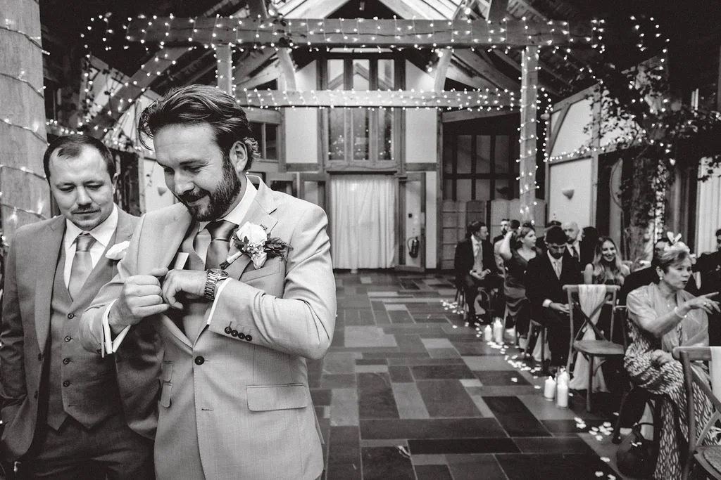 A man in a suit with a boutonniere adjusting his watch at a wedding reception, with another man beside him and guests seated in the background, decorated with string lights and candles, in a rustic indoor venue.