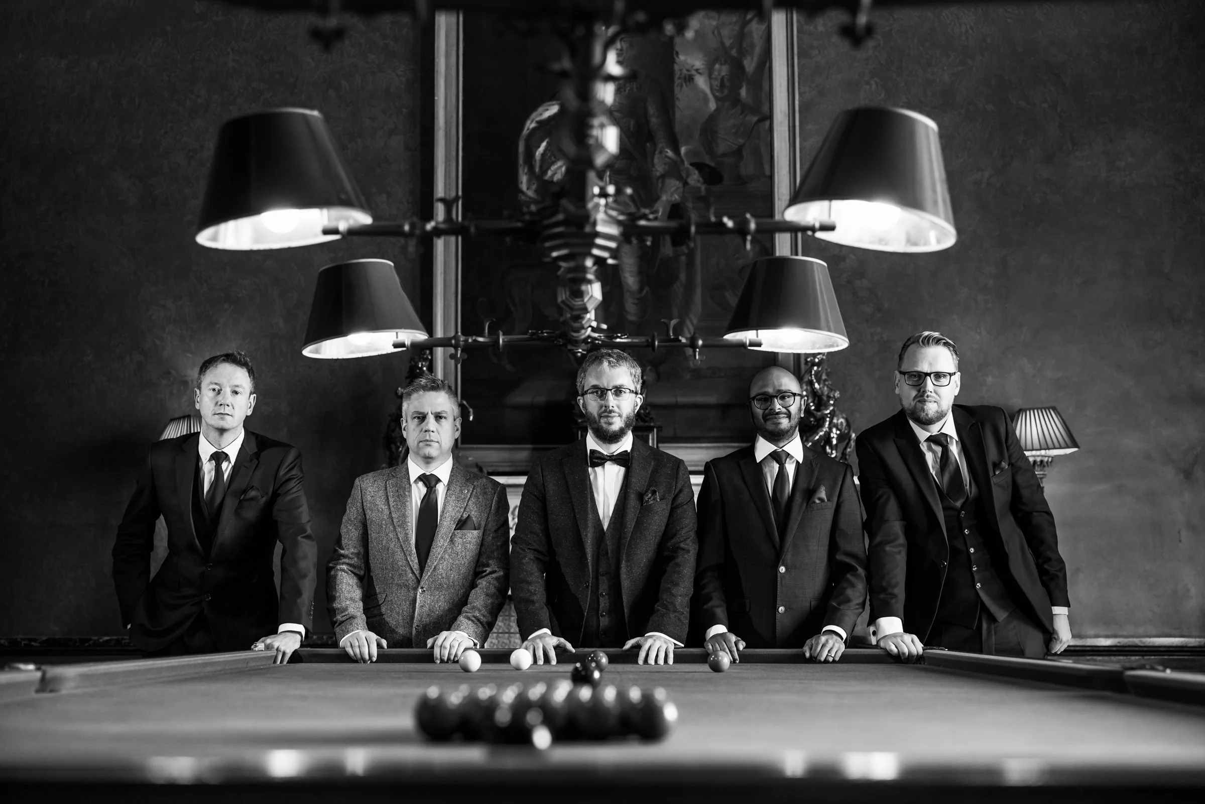 Black and white photo of five men dressed in formal suits standing behind a billiards table, with a large chandelier hanging above and a framed painting on the wall.