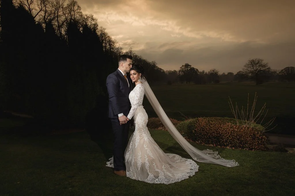 A bride and groom standing together outdoors at sunset, with the bride wearing a long white wedding gown and veil, and the groom in a dark suit, holding hands and gazing at each other.