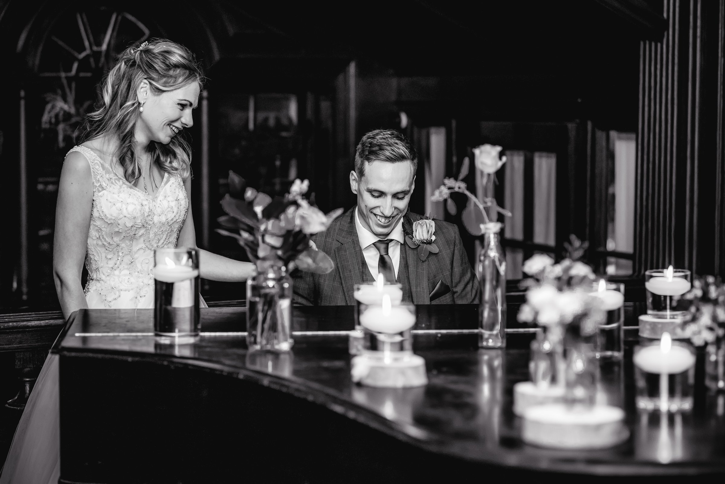 A black and white photo of a wedding scene with a bride and groom. The bride is standing next to a table with flowers and candles, smiling as the groom sits and looks down, also smiling.
