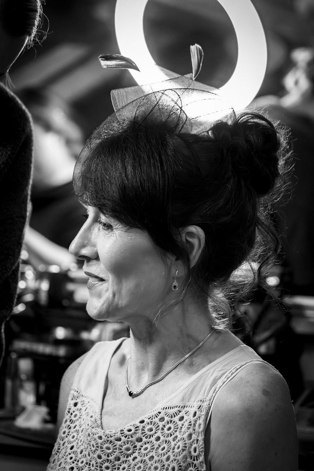 A woman in profile with dark hair, wearing a lace top, earrings, and a necklace, is smiling. The background is blurred, with circular lighting and other people partially visible, suggesting a social or celebration event.