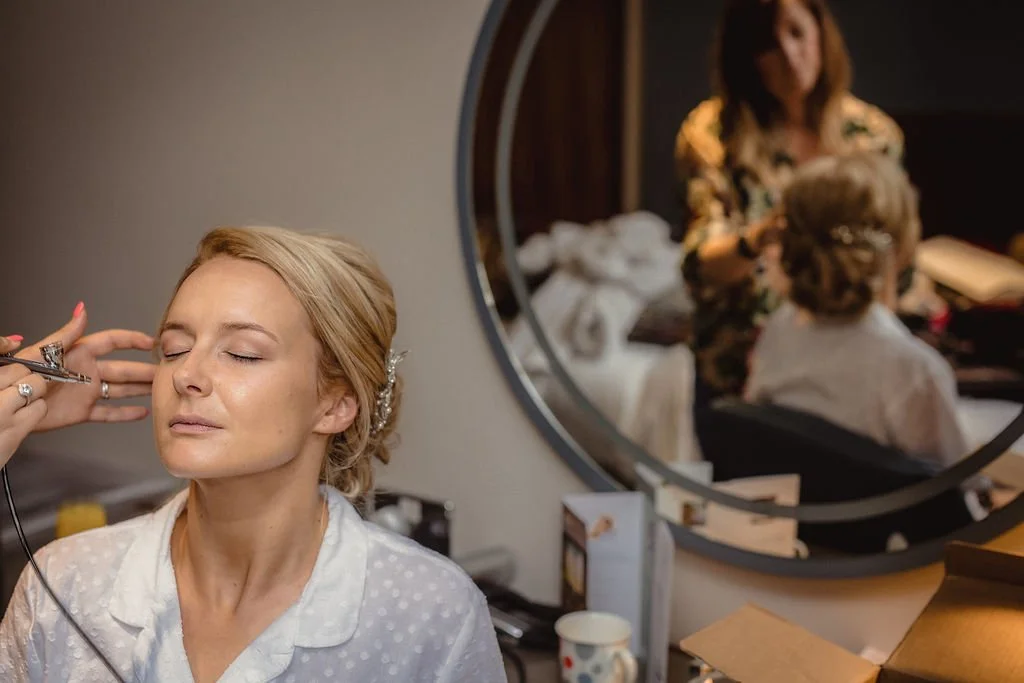 A woman with blonde hair getting her makeup done in front of a mirror, with another woman and a child reflected in the mirror.