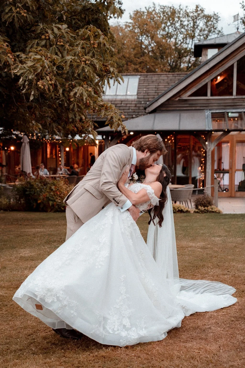 A bride and groom sharing a kiss outdoors during sunset, with the bride in a white wedding gown and veil, and the groom in a beige suit, in front of a rustic building decorated with string lights.