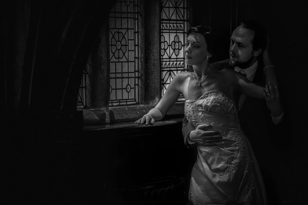 A black and white photograph of a woman in a wedding dress leaning against a window with stained glass, while a man in formal attire stands behind her, gently holding her waist.