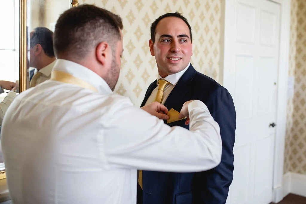 Two men dressing up in suits, one adjusting the other's tie in a room with patterned wallpaper and a mirror.