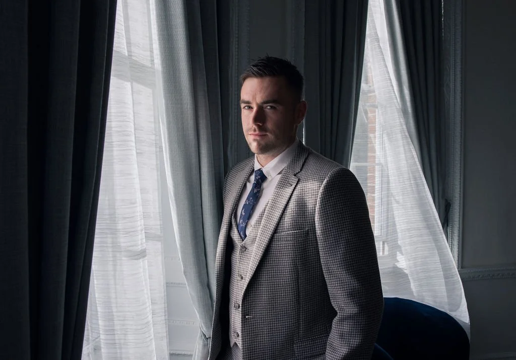 A man in a gray checked suit standing by a window with sheer curtains, looking at the camera.