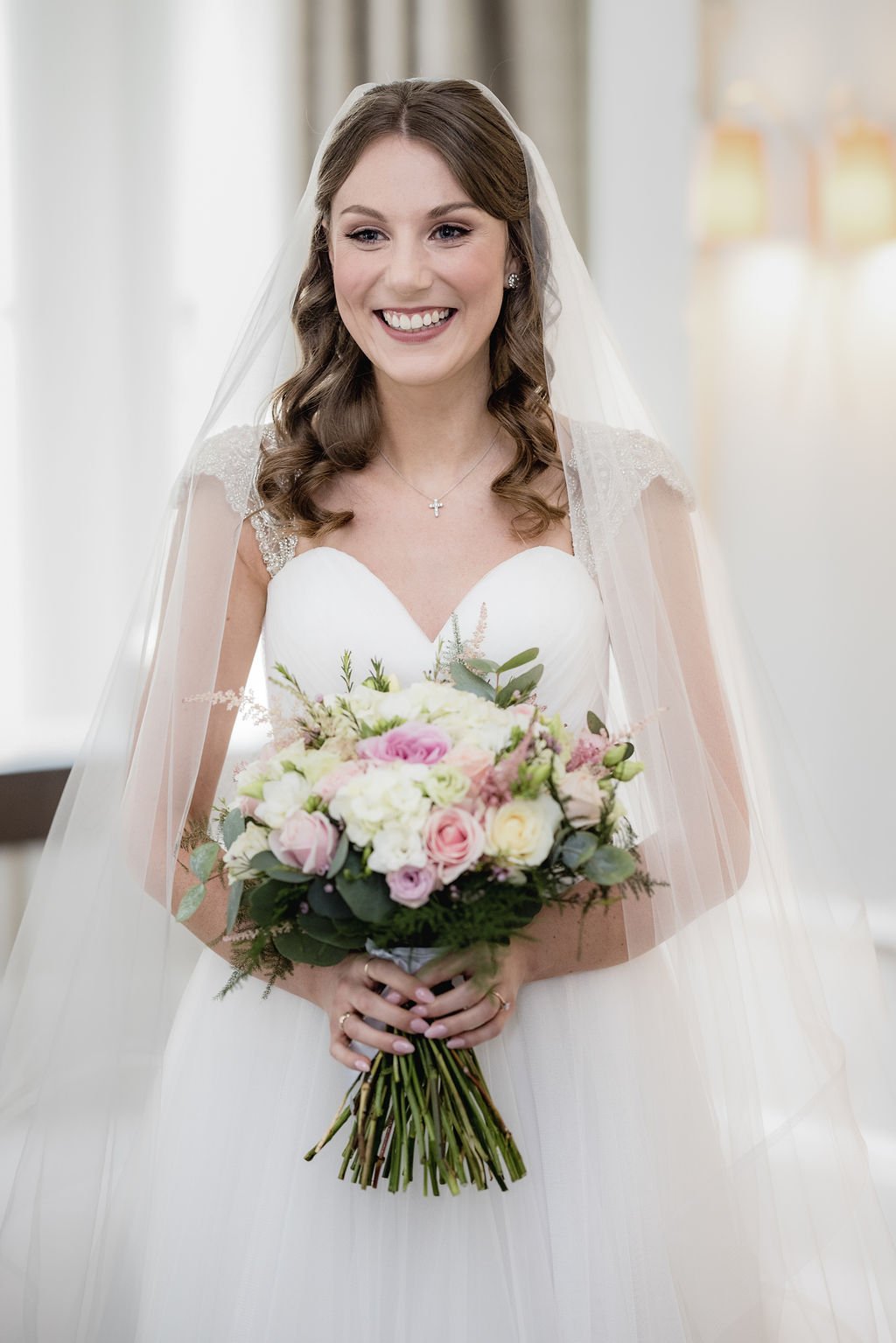 A smiling bride with wavy brown hair in a white wedding dress holding a bouquet of pink and white flowers, wearing a veil and a cross necklace.