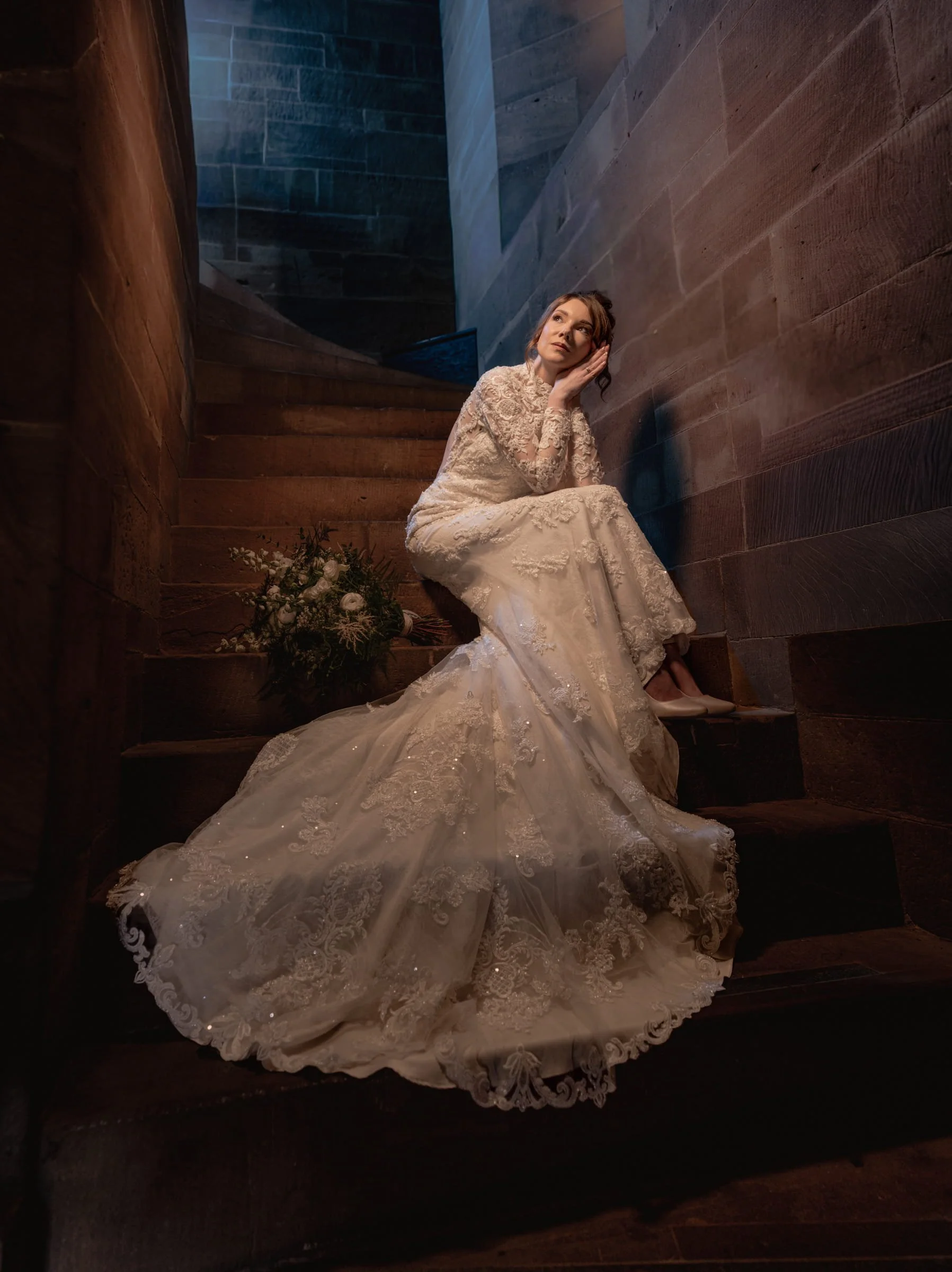 A woman in a white lace wedding dress sitting on a wooden staircase, with her head resting against the wall and gazing thoughtfully upward. A bouquet of flowers is placed at her feet.