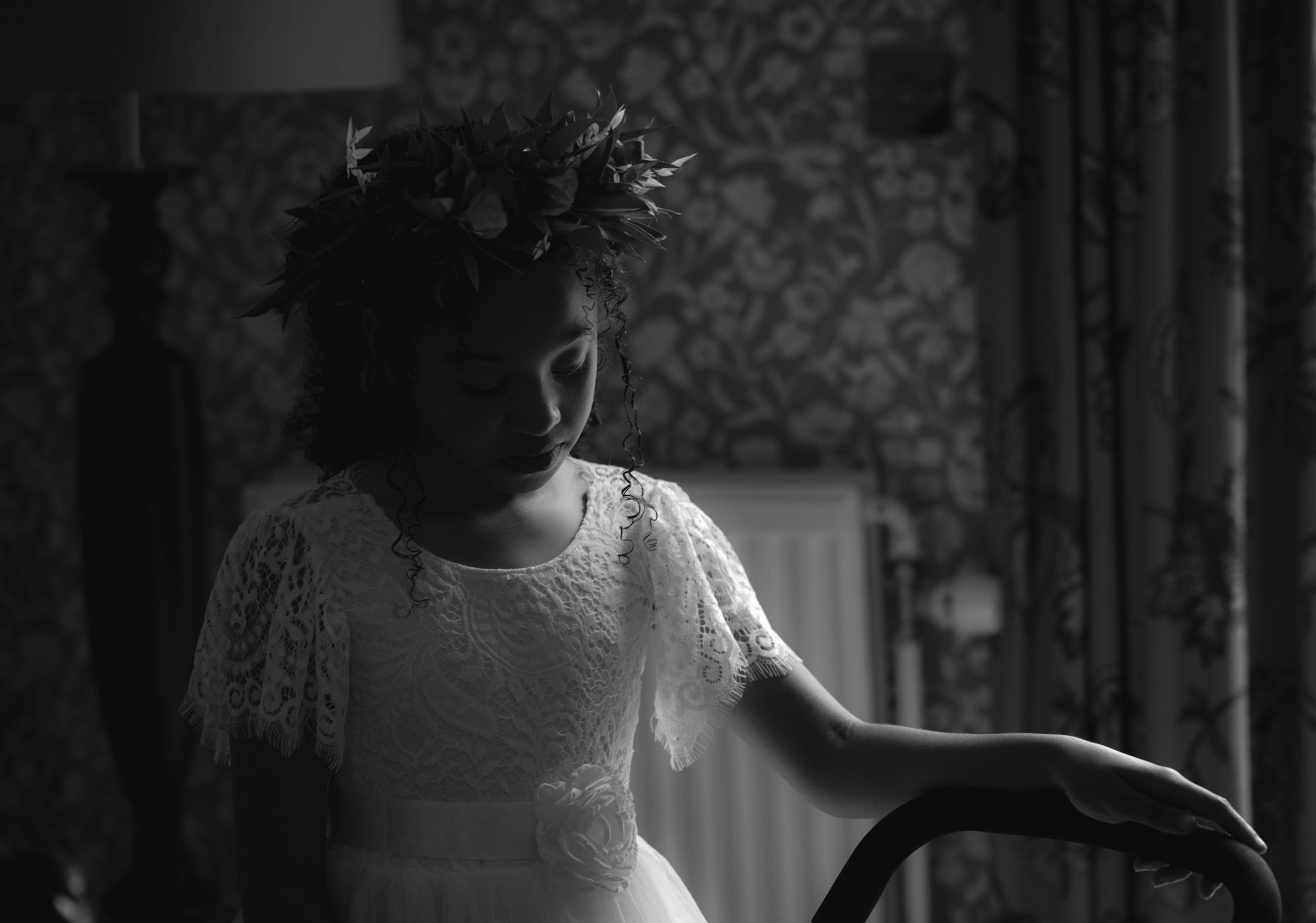 A young girl wearing a lace dress and a floral crown, standing indoors with her eyes closed and hand on a chair.