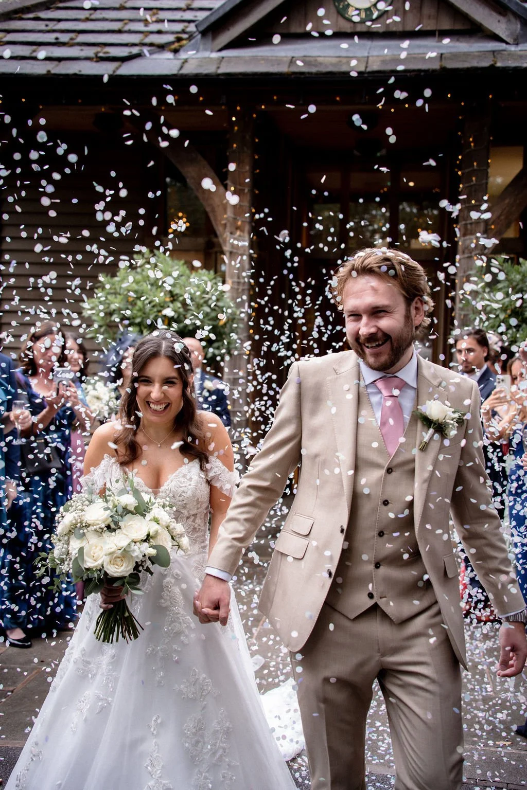 A newlywed couple holding hands and smiling as confetti falls around them, with guests in the background celebrating outside a rustic building.