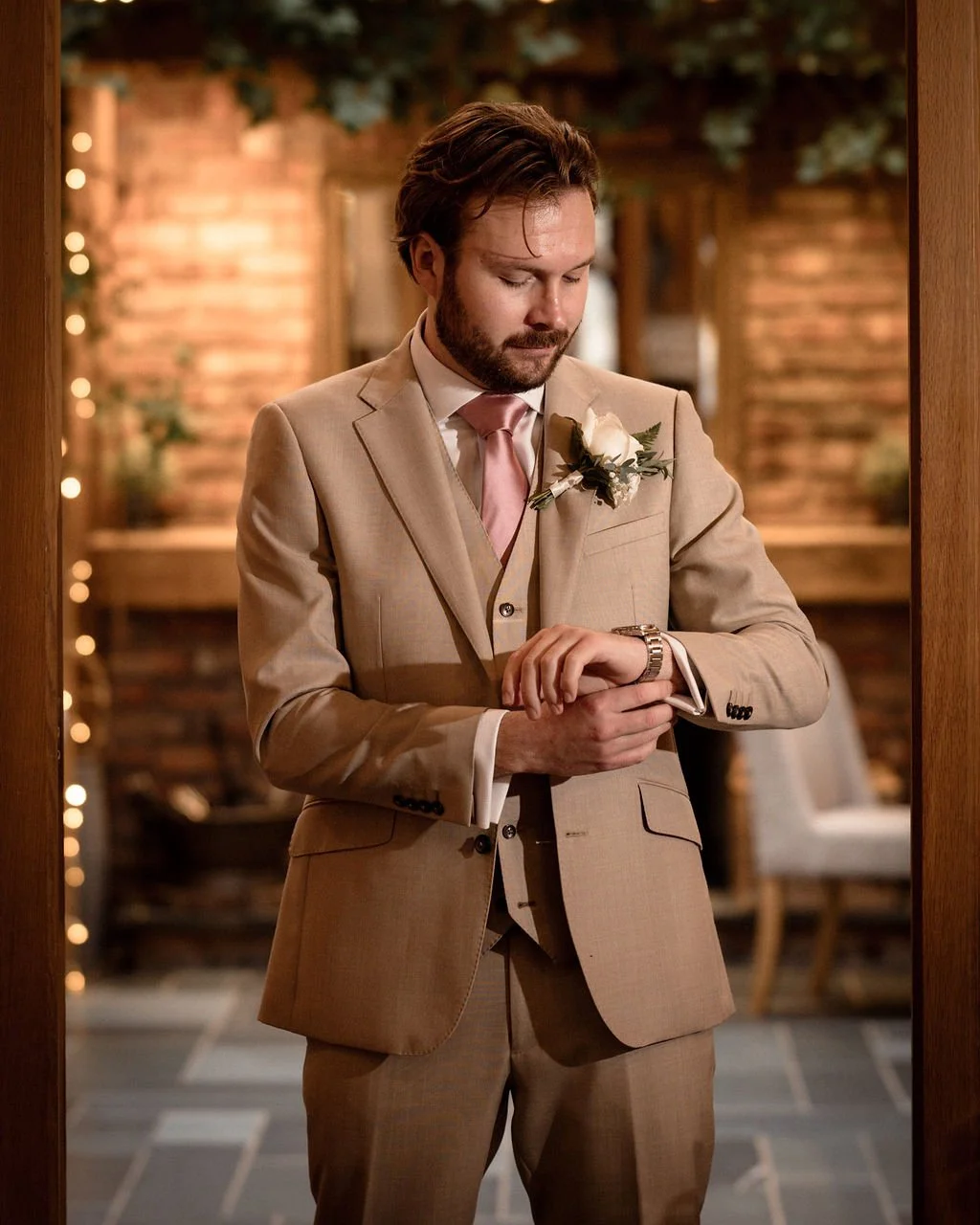 A man in a beige suit with a pink tie and a white rose boutonniere looks at his wristwatch.