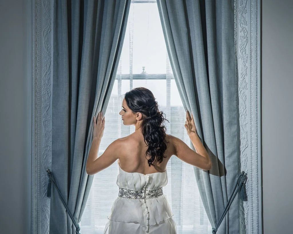 A woman in a white strapless dress with a bejeweled waist sits by a window, holding open blue curtains and looking outside.