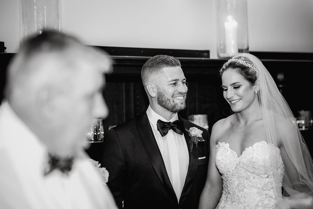 A bride and groom smiling at each other during their wedding ceremony, with a blurred person in the foreground and candles in the background.
