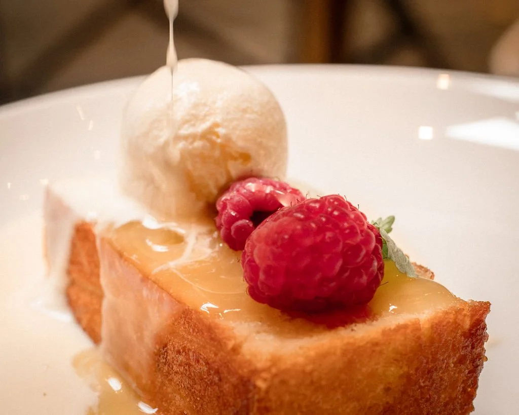 A dessert with a slice of cake topped with raspberries, an ice cream ball, and syrup being poured over it.