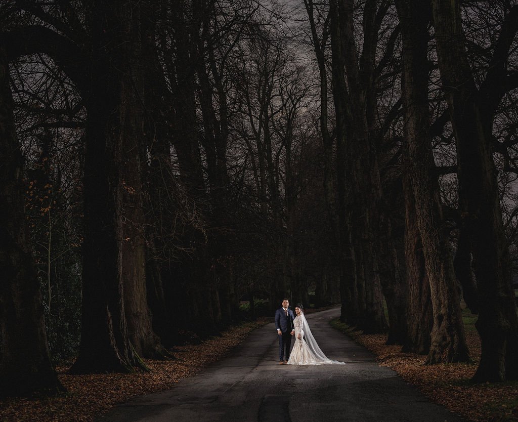 A bride and groom in wedding attire holding hands on a dark, tree-lined road at night.