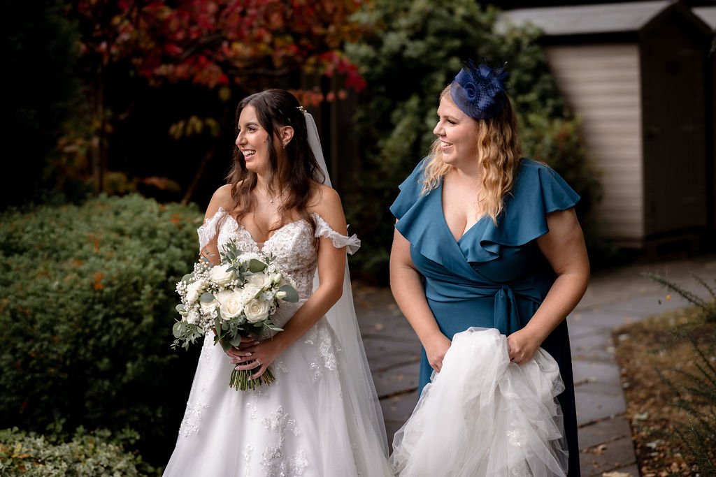 A bride in a white wedding dress holding a bouquet and smiling, standing outside next to a woman in a blue dress wearing a matching hat, both smiling and enjoying a moment together.
