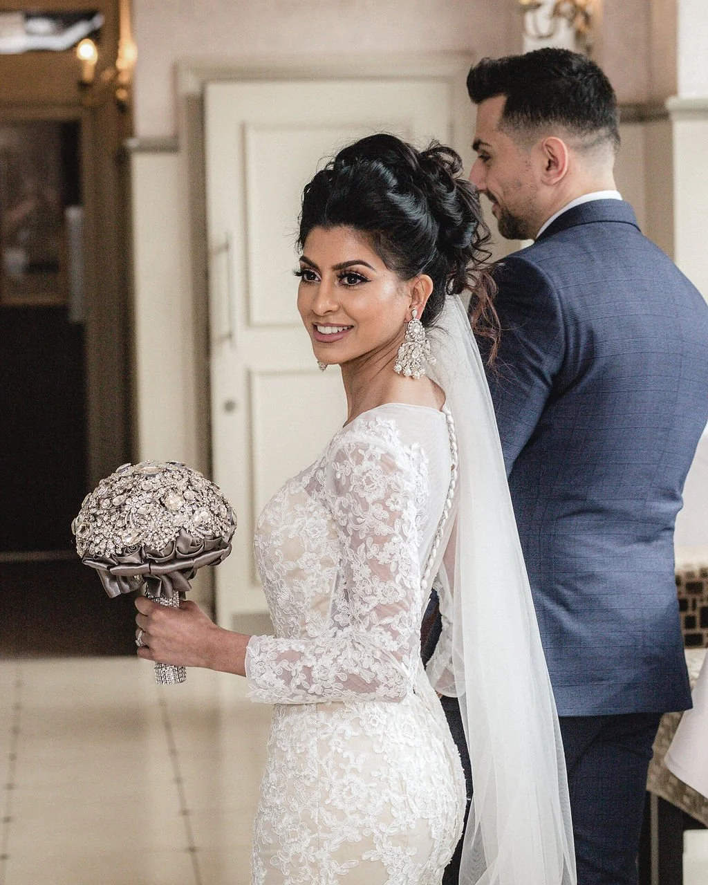 A bride in a white lace wedding dress with long sleeves holding a bouquet, smiling, with a groom in a navy suit standing behind her, in an indoor setting.