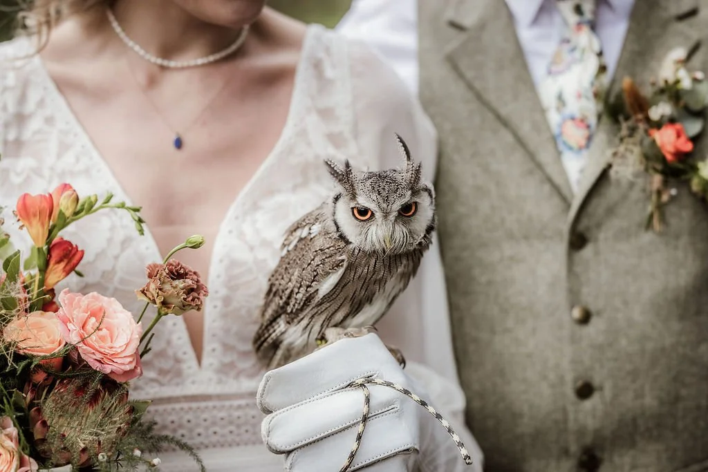 A woman holding an owl on her gloved hand, standing next to a man in a gray suit and floral shirt, with a bouquet of pink and peach flowers in front.