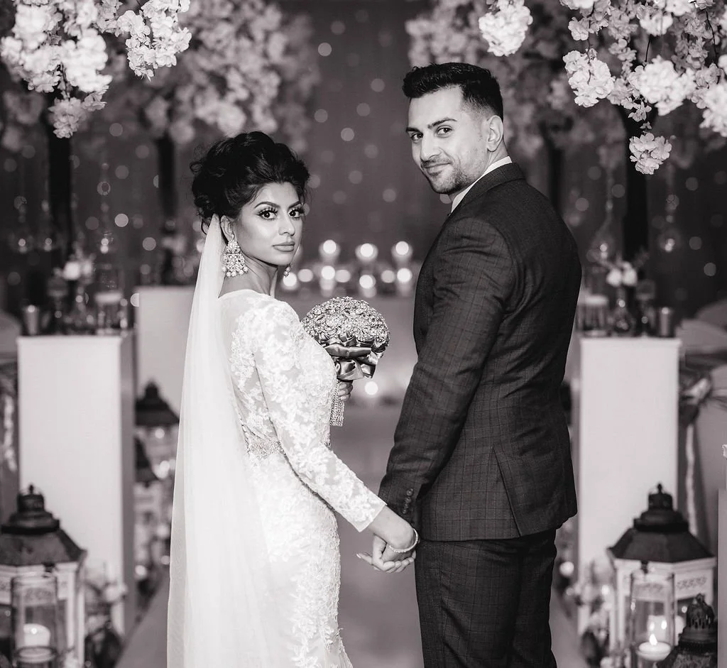 A black and white photo of a bride and groom standing and holding hands at their wedding reception, with floral and decorative backdrop.