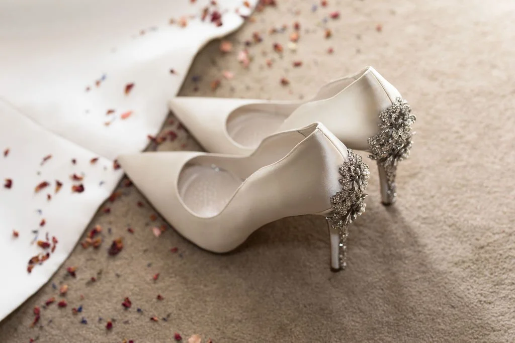 White high-heeled wedding shoes with jeweled embellishments on the heels, resting on a carpet near a white fabric with scattered flower petals.