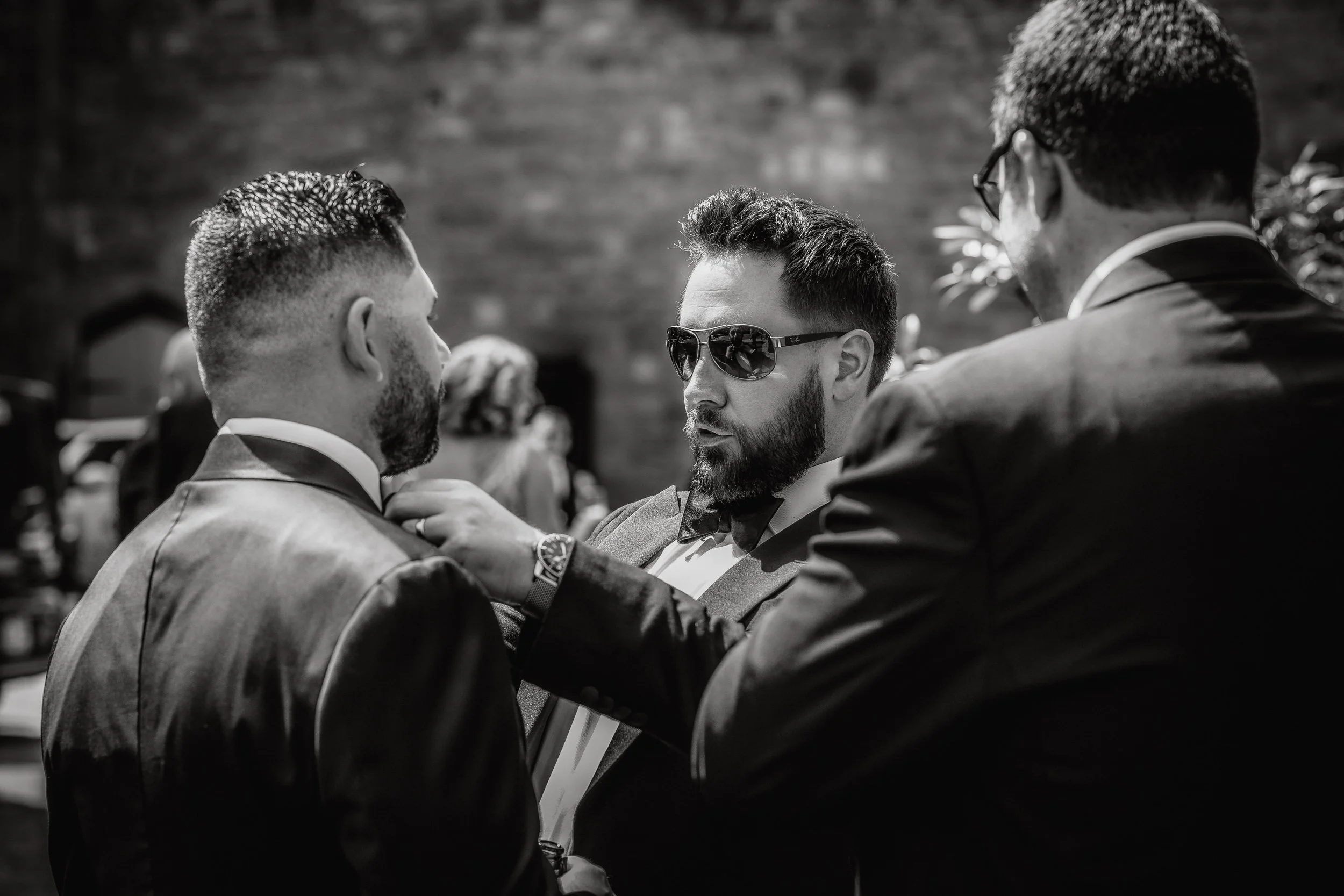 Three men in tuxedos, two of them touching the third man's tuxedo lapel, one of them wearing sunglasses, in a formal setting.