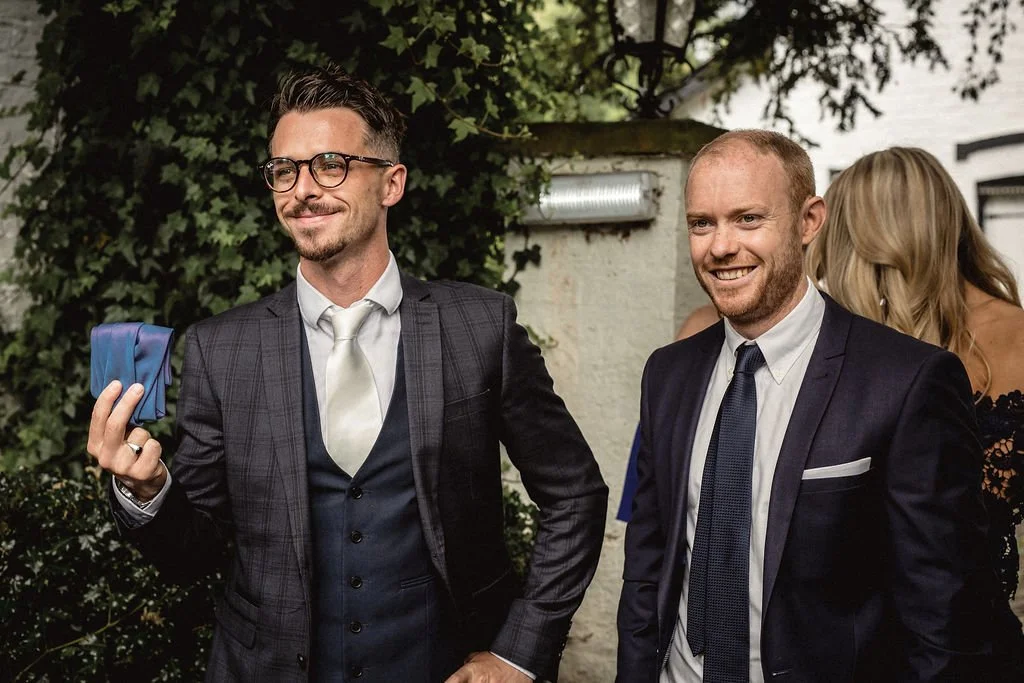 Two men in suits smiling at an outdoor event, with greenery and a woman in a black dress in the background.