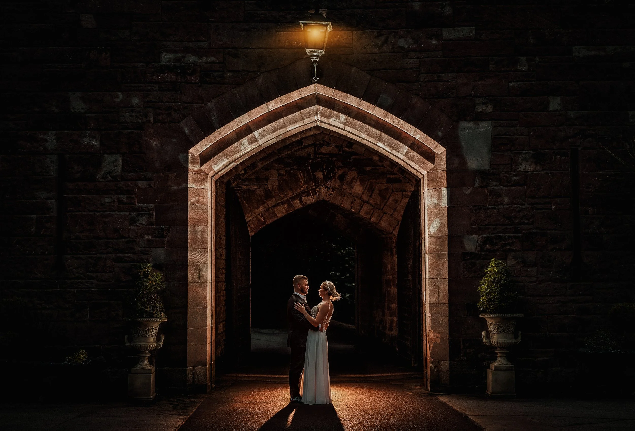 Bride and groom standing close under an arched brick gate at night, illuminated by a warm lamp above, with potted plants on either side.