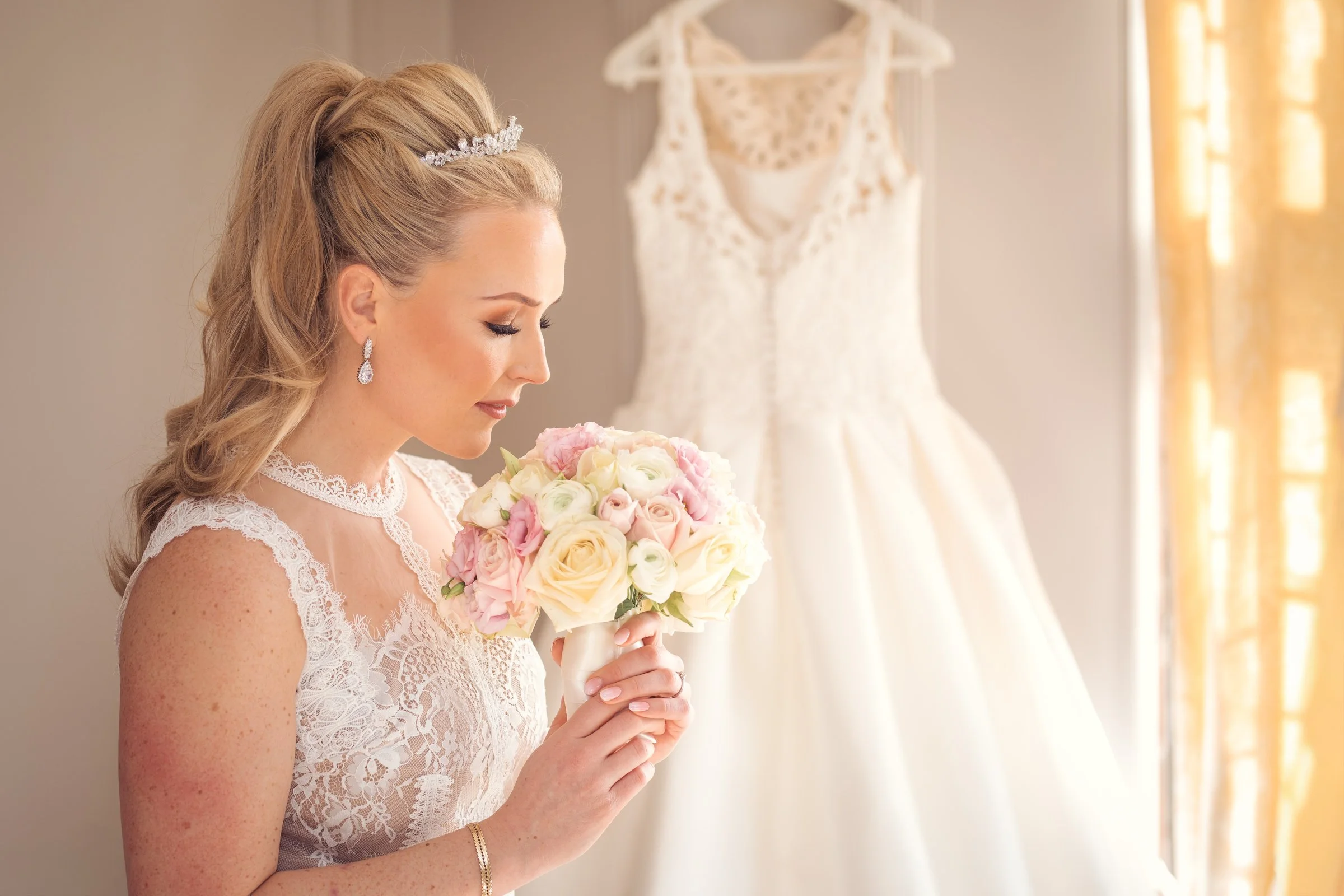 A bride in a lace dress holding a bouquet of pink and white roses, standing in front of a white wedding gown hung on a wall.