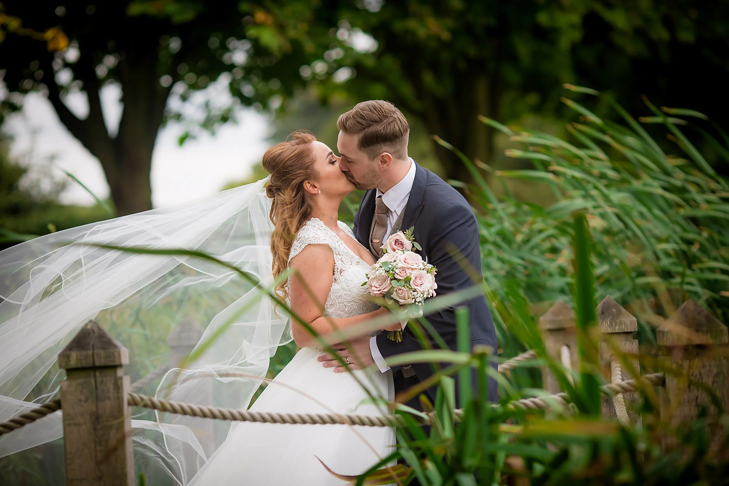 A bride and groom share a kiss outdoors on their wedding day, with the bride holding a pink and white bouquet, and greenery and trees in the background.