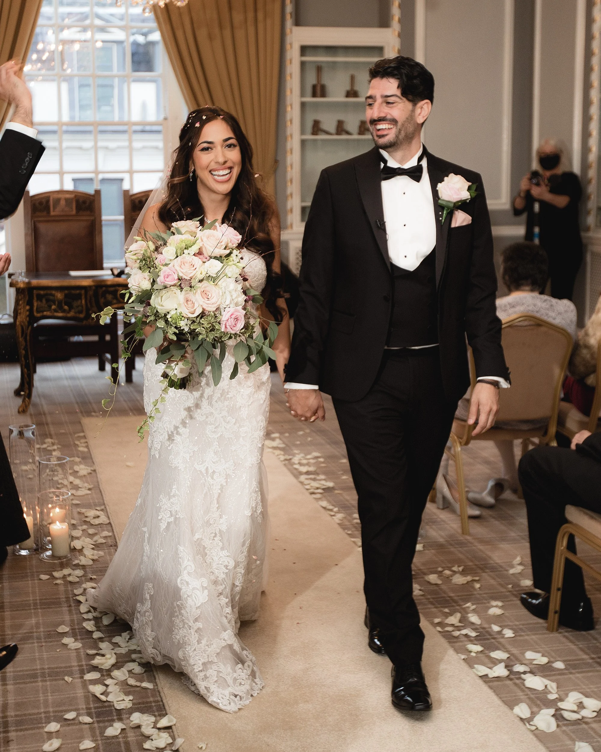 A newlywed couple walking down the aisle during their wedding ceremony in an elegant indoor venue, surrounded by seated guests, with floral arrangements and lit candles.