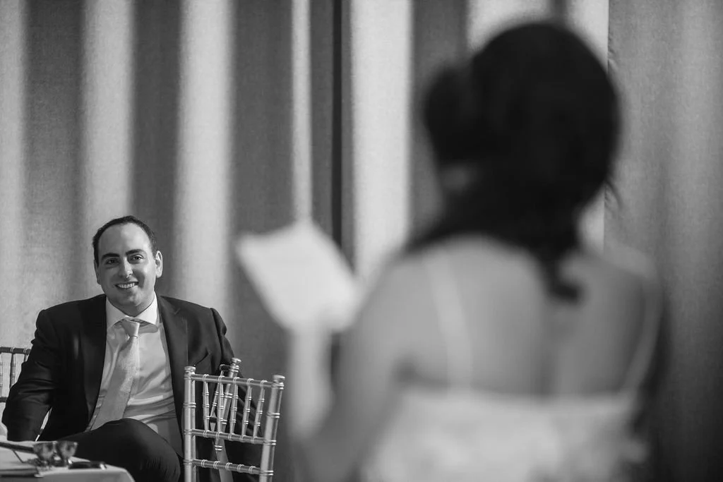 A man in a suit sitting at a table and smiling, seen from behind a woman whose face is blurred in the foreground.