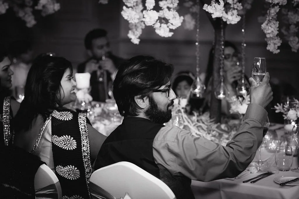 People sitting at a table during a celebration, some holding drinks, with floral decorations hanging above.
