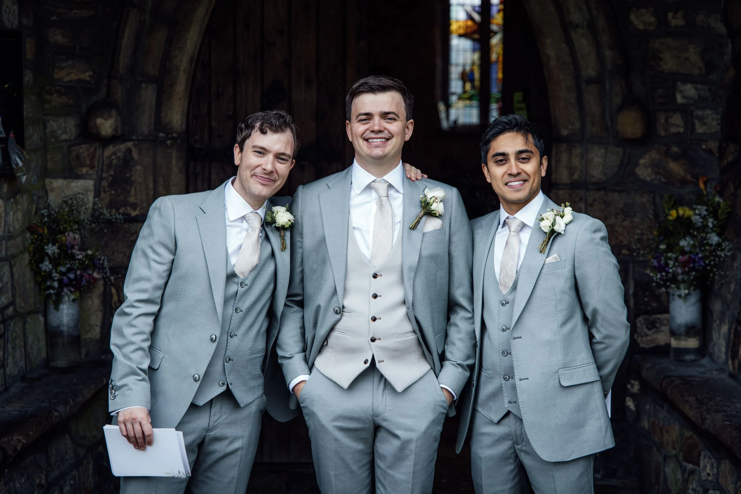 Three men in suits smiling at a wedding ceremony inside a stone church, with stained glass windows in the background. The groom is in the middle, with two groomsmen on each side.