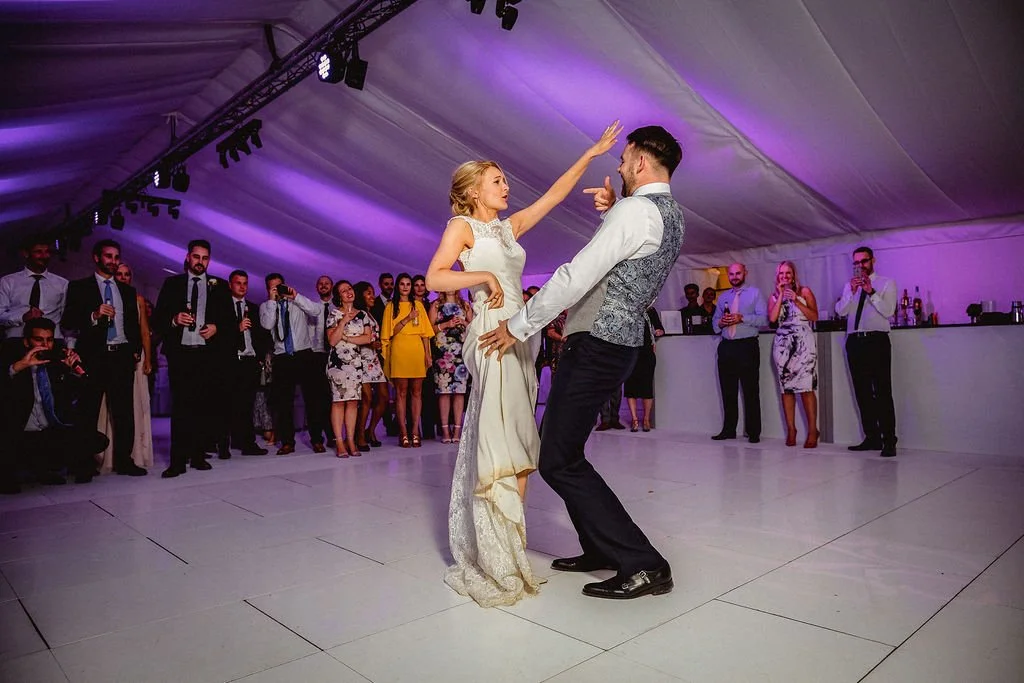 A bride and groom dancing together at their wedding reception under purple lighting, with guests watching and taking photos in the background.