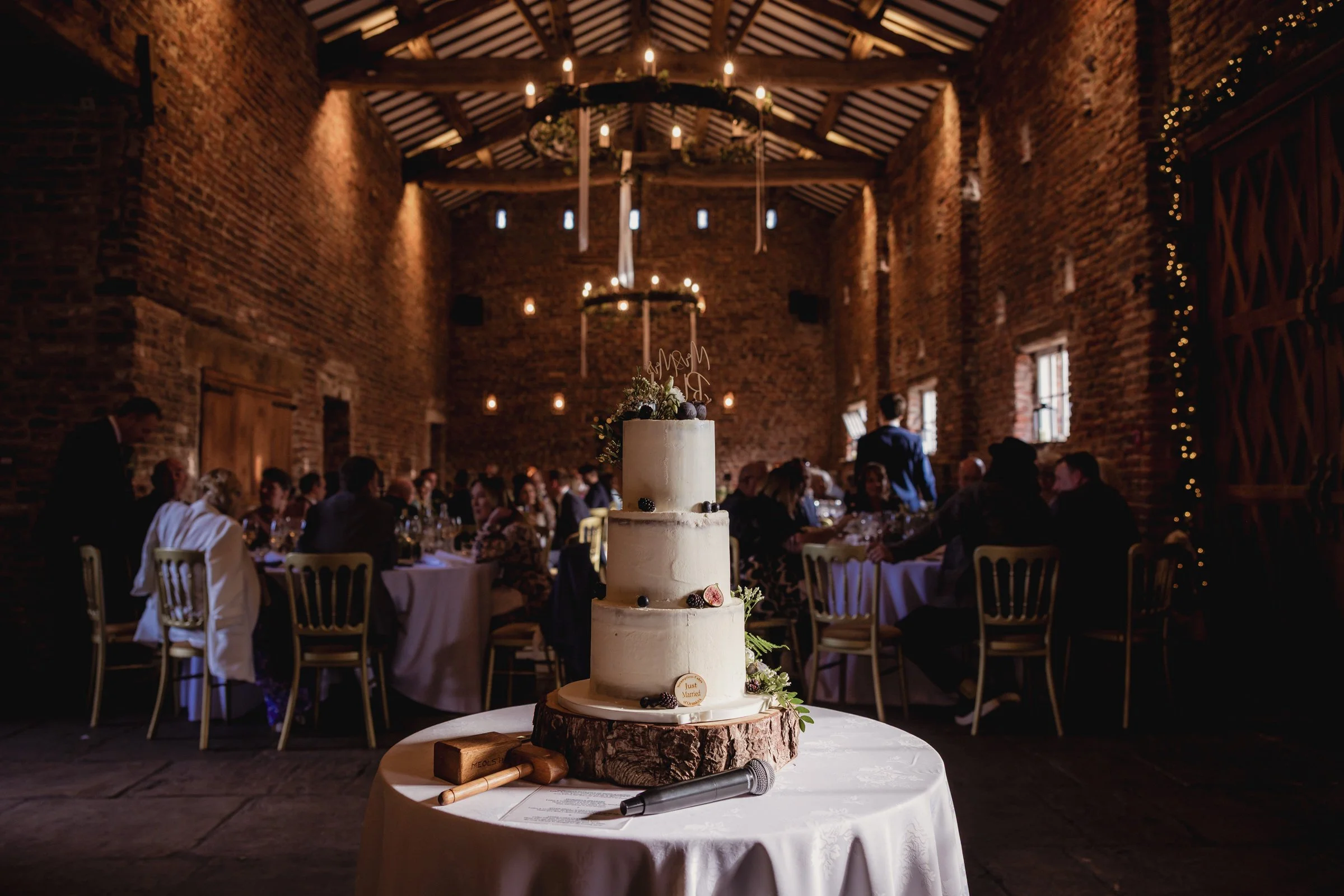 A multi-tiered wedding cake decorated with berries and flowers, placed on a wooden slab on a round table covered with a white tablecloth, in a rustic barn with brick walls and chandeliers, during a wedding celebration.