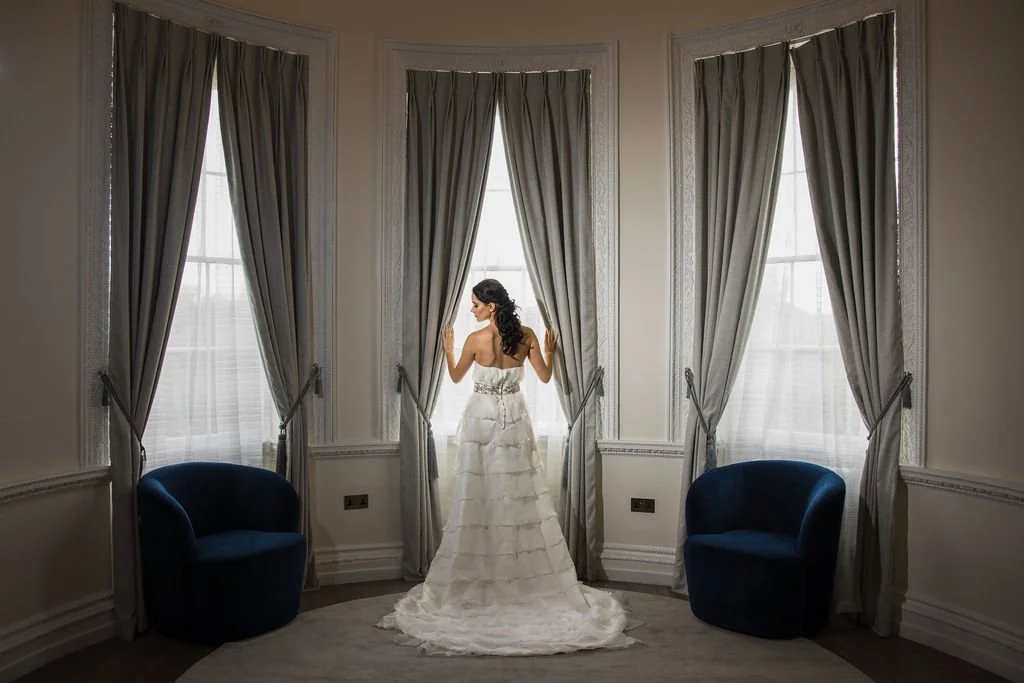 A bride in a white wedding dress stands between three large windows with tall curtains, flanked by two blue chairs in a formal room.