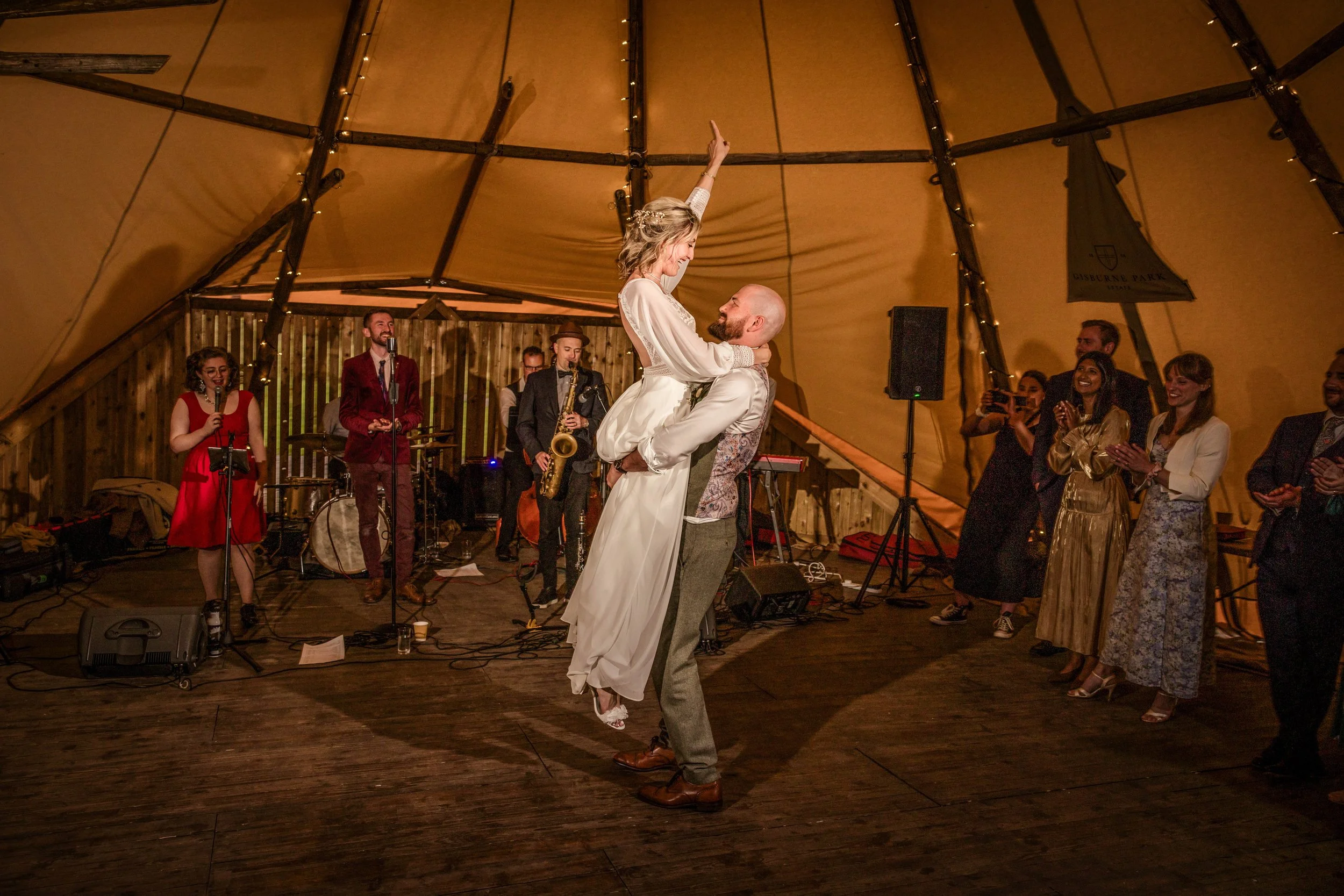 A couple dancing at a wedding reception, with the groom lifting the bride. The bride is wearing a white dress, and the groom is dressed in a vest and trousers. A live band is playing in the background, and guests are watching and clapping.