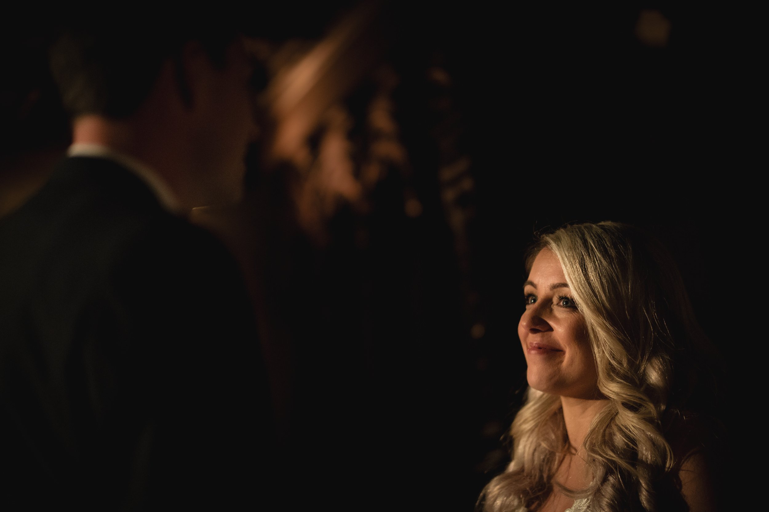 A woman with long, blonde hair smiling softly as she looks at a man in a dark suit in a dimly lit setting.