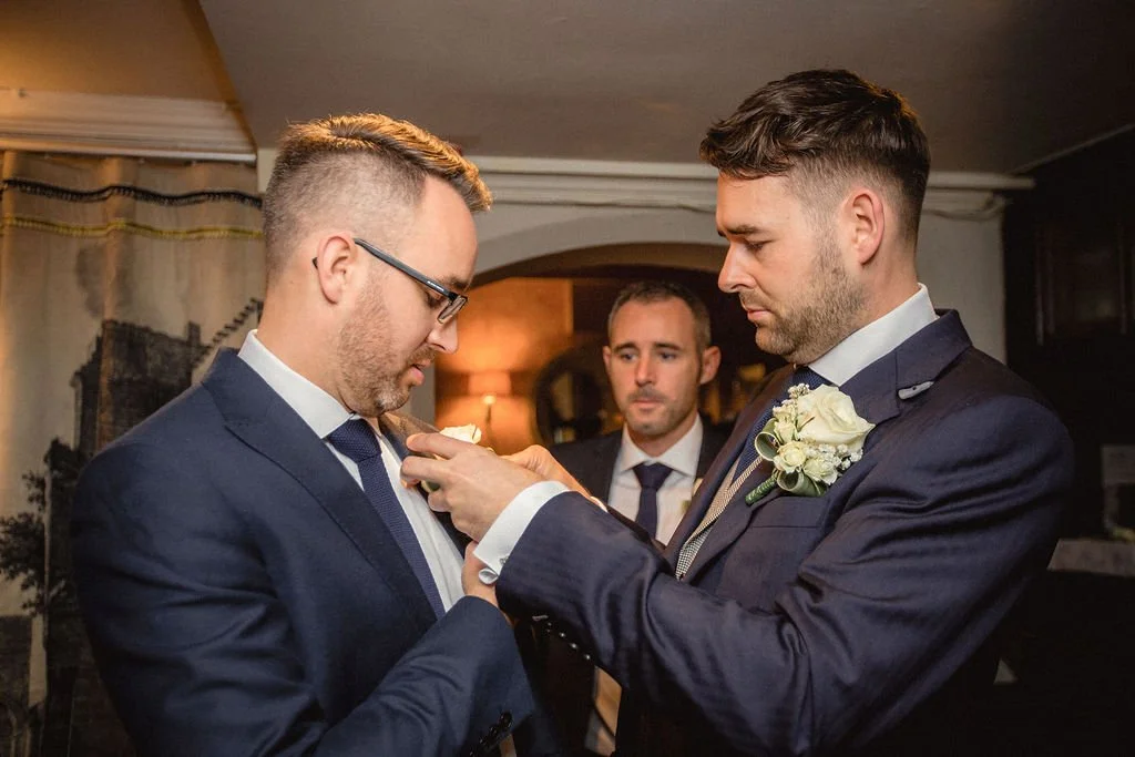 Two men in suits with boutonnières are exchanging or adjusting a pin or badge, while a third man looks on in a warmly lit room.