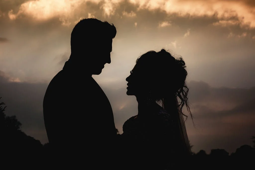Silhouette of a man and woman facing each other during sunset or sunrise, with a cloudy sky in the background.
