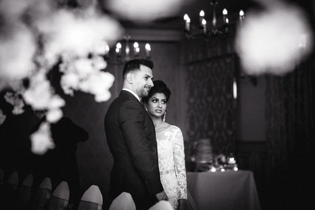 A black and white photo of a wedding reception with a groom and bride standing together. The groom is in a dark suit, smiling, and the bride is in a lace wedding dress, looking worried or anxious. There are chairs at the bottom and floral decorations