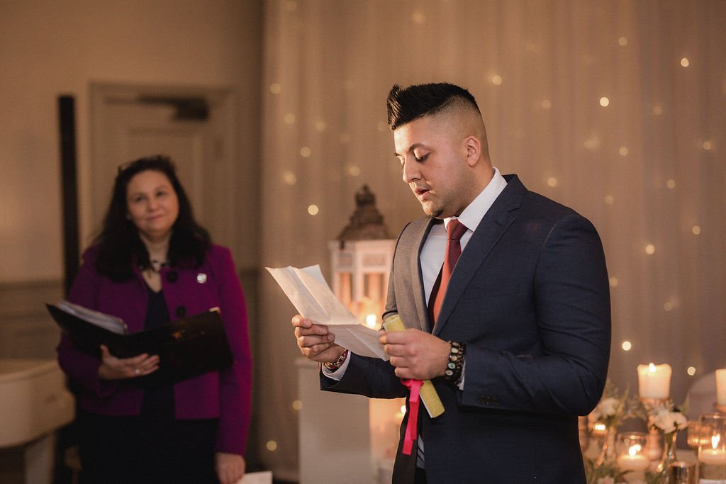A man in a dark suit and red tie standing in front of a beige curtain with small lights, reading from a paper. A woman in a purple jacket holding a black folder observes him.